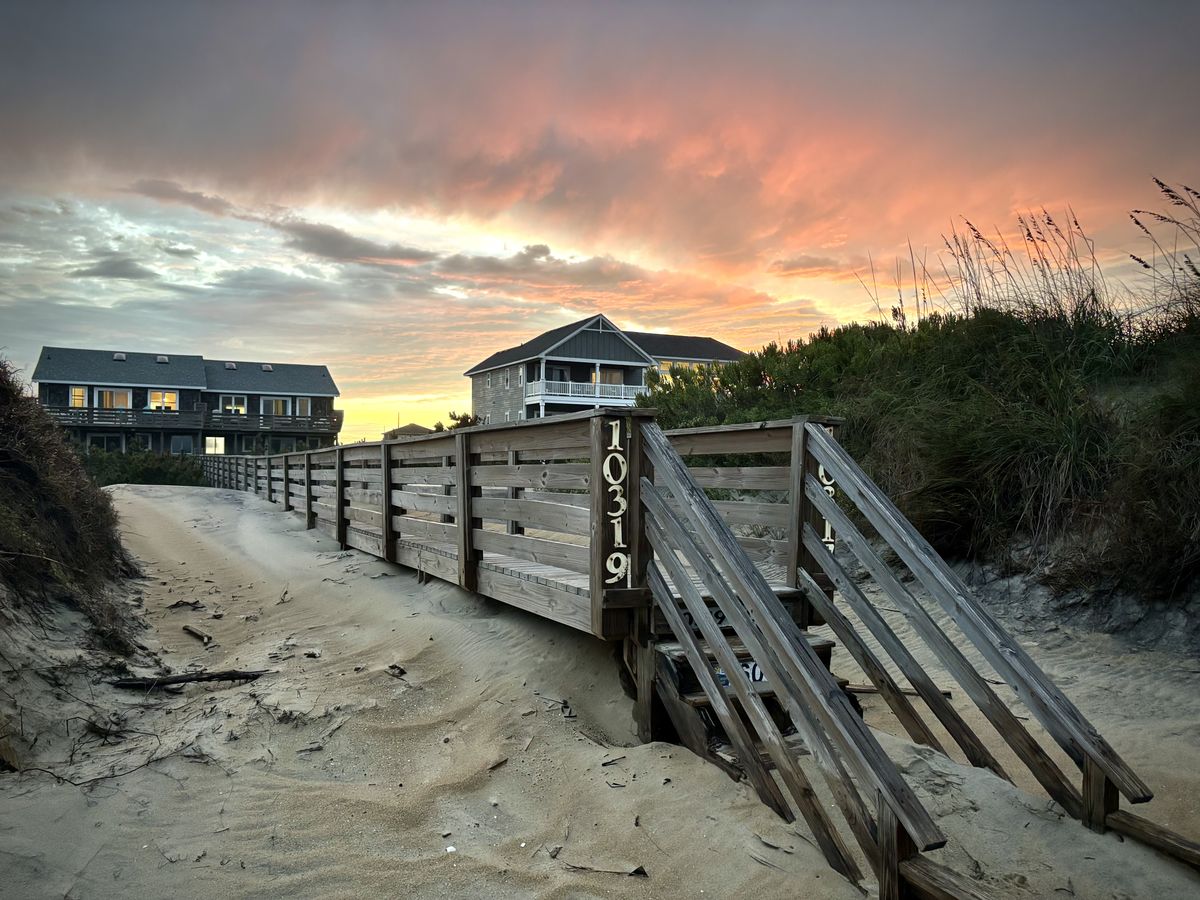 A beach house walkway, with a sand dune in the foreground and house + sunset in the background