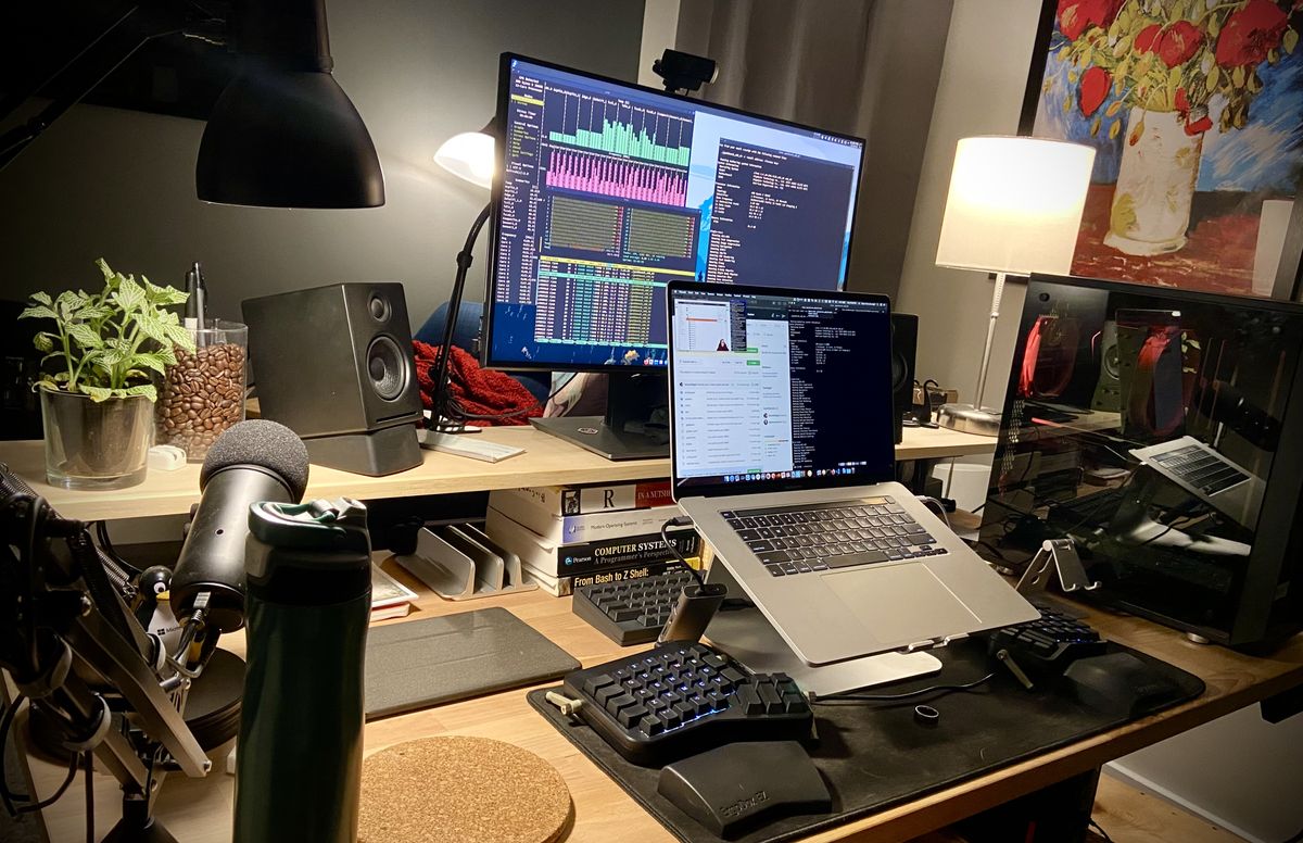 A MacBook Pro on a stand, between a split keyboard, with a desktop to the side of it and monitor behind.
