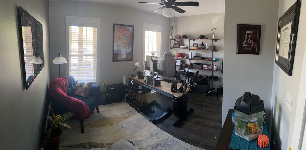 A wide angle shot from the entrance of a home office, with a wooden desk in the center, wood+pipe shelf behind it, and cozy rug and armchair setup in the front.