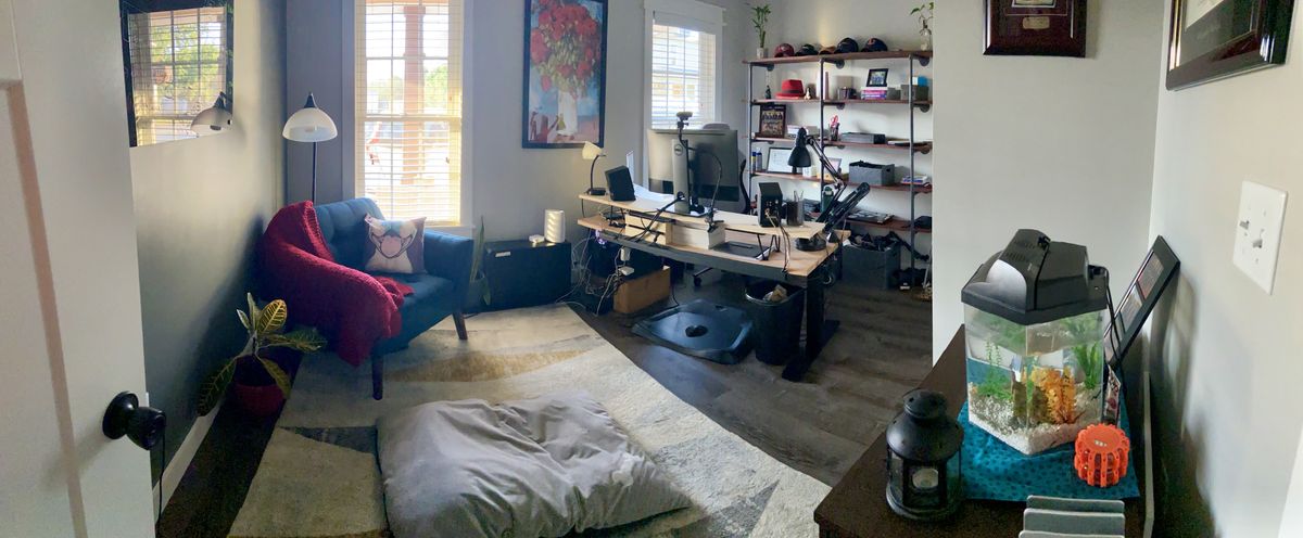 A wide angle shot from the entrance of a home office, with a wooden desk in the center, wood+pipe shelf behind it, and cozy rug and armchair setup in the front.