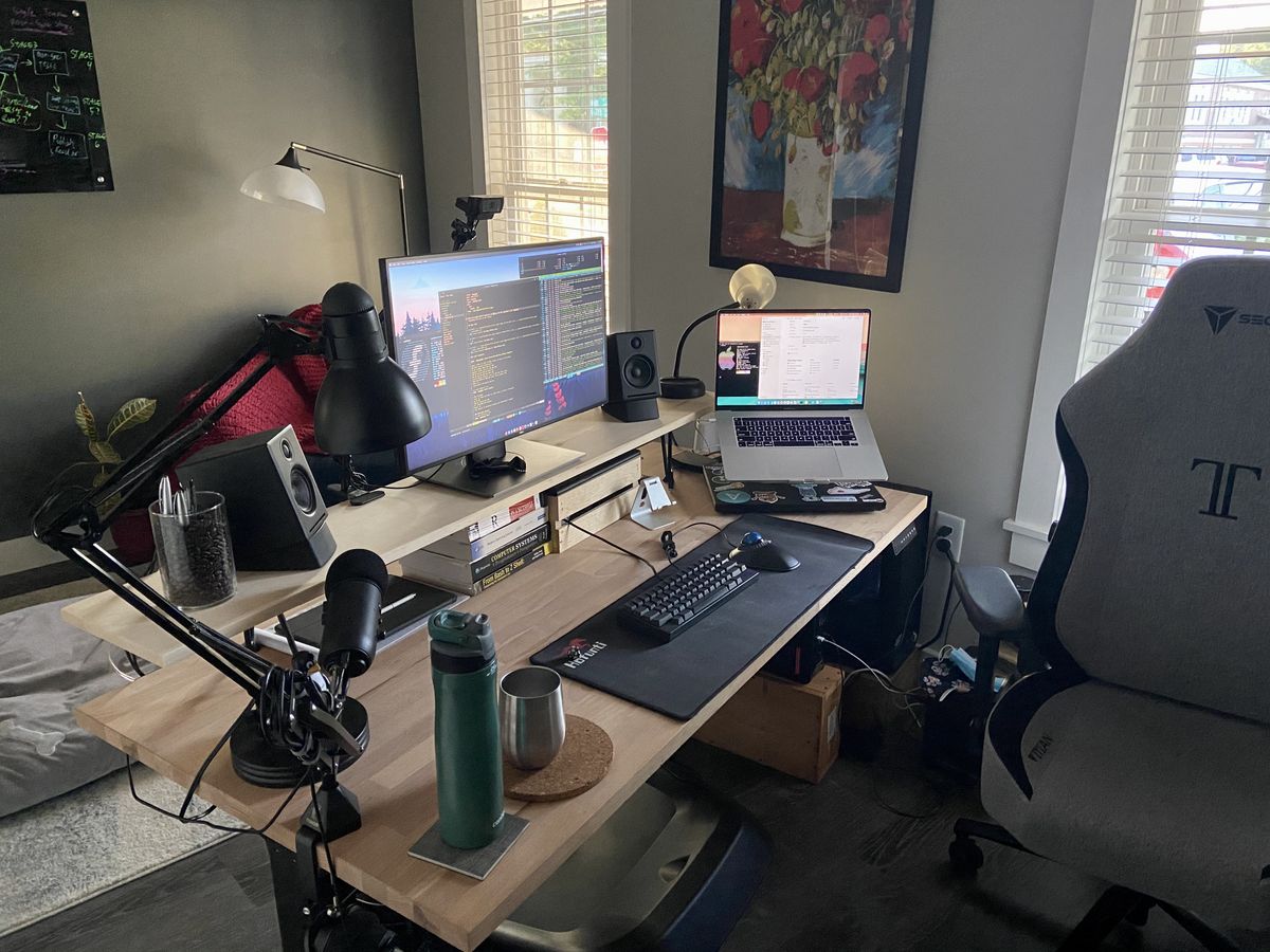 A wooden computer desk setup, with a monitor on a riser shelf, a macbook stand, and a chair