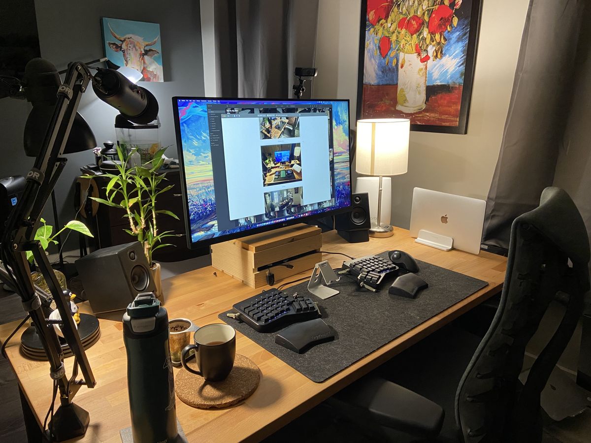 A wooden computer desk with a large monitor, split keyboard, warm lights, and some plants
