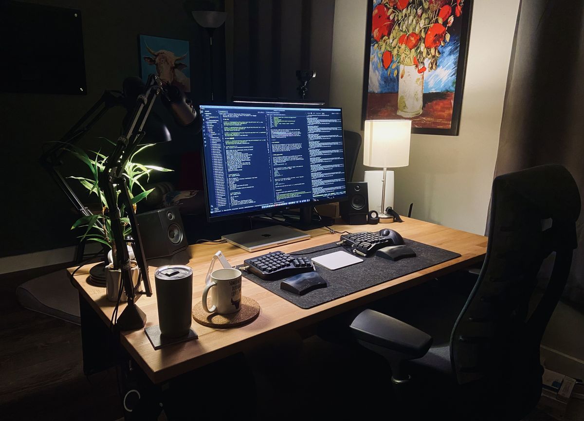 A wooden desk lit in a home office with a large monitor and split keyboard