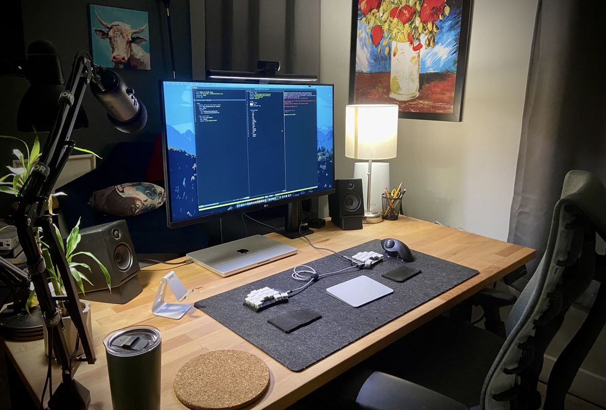 A lamp-lit wooden desk in a dark home office, with a large monitor and a small split keyboard.