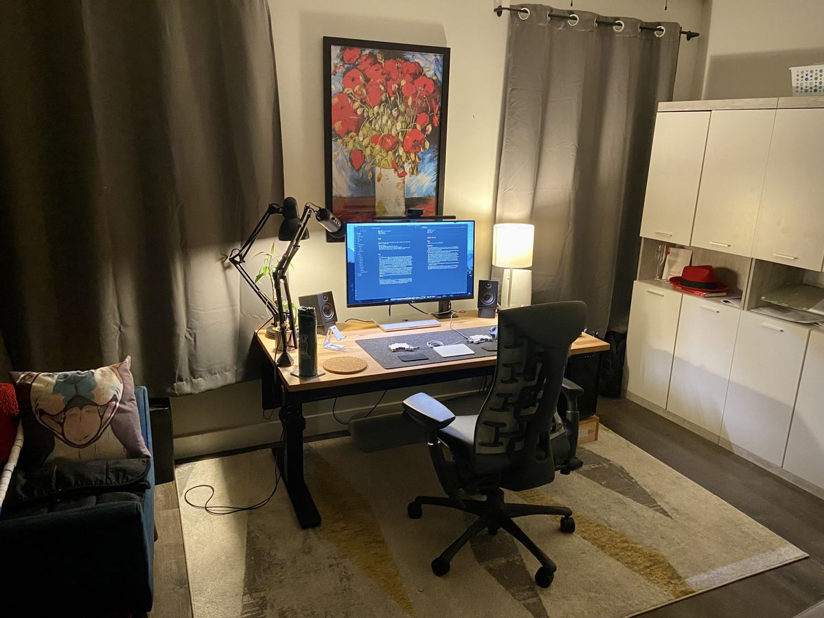 A lamp lit wooden desk between two curtained windows with a red flower painting behind it. A large monitor, small split keyboard, and speakers are also on the desk. The desk and office chair are on a rug, with shelves to the right side of the room and a cozy arm chair to the left