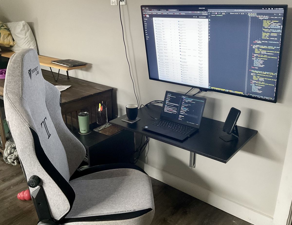 A laptop on a small folding wall desk, with a giant monitor wall mounted above it