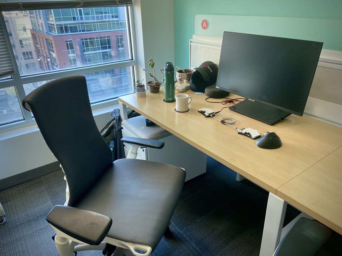 An end desk inside an office, with a window behind it. A mounted monitor, closed thinkpad, split keyboard, trackball, coffee, and some plants are on the desk