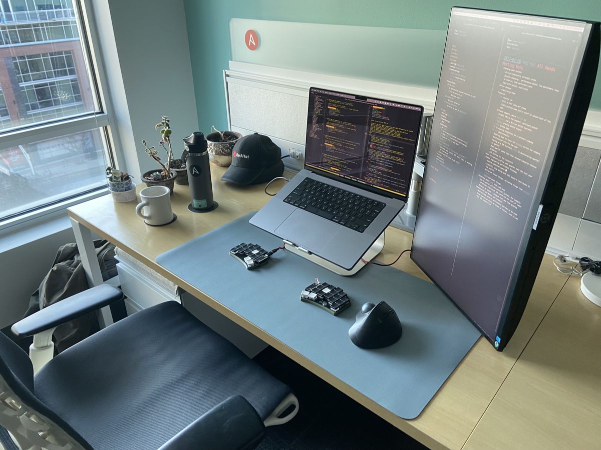 An end desk inside an office, with a window behind it. A MacBook on a stand, with a mounted portrait monitor on the right side it. A split keyboard, trackball, coffee, and some plants are on the desk