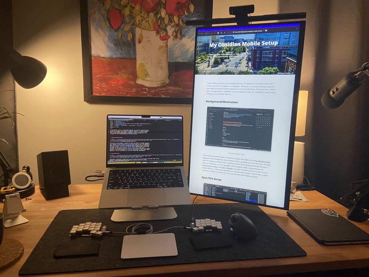 A lamp lit wooden desk with a small macbook pro on a stand, split keyboard, and large monitor in portrait mode to the right of it